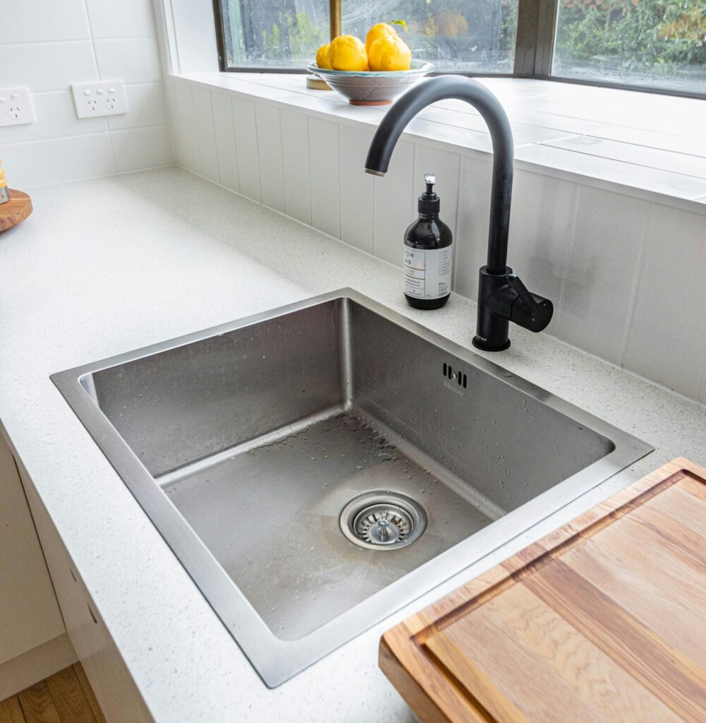 Kitchen sink in a Pennsylvania home. Single stainless steel sink with matte black faucet surrounded by white stone countertop and wood cutting board  to the right.